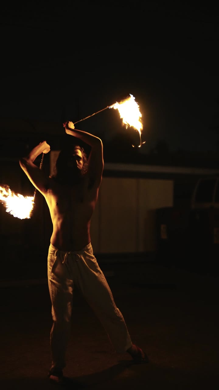 Shirtless spinner with double poi trailing flame across the frame